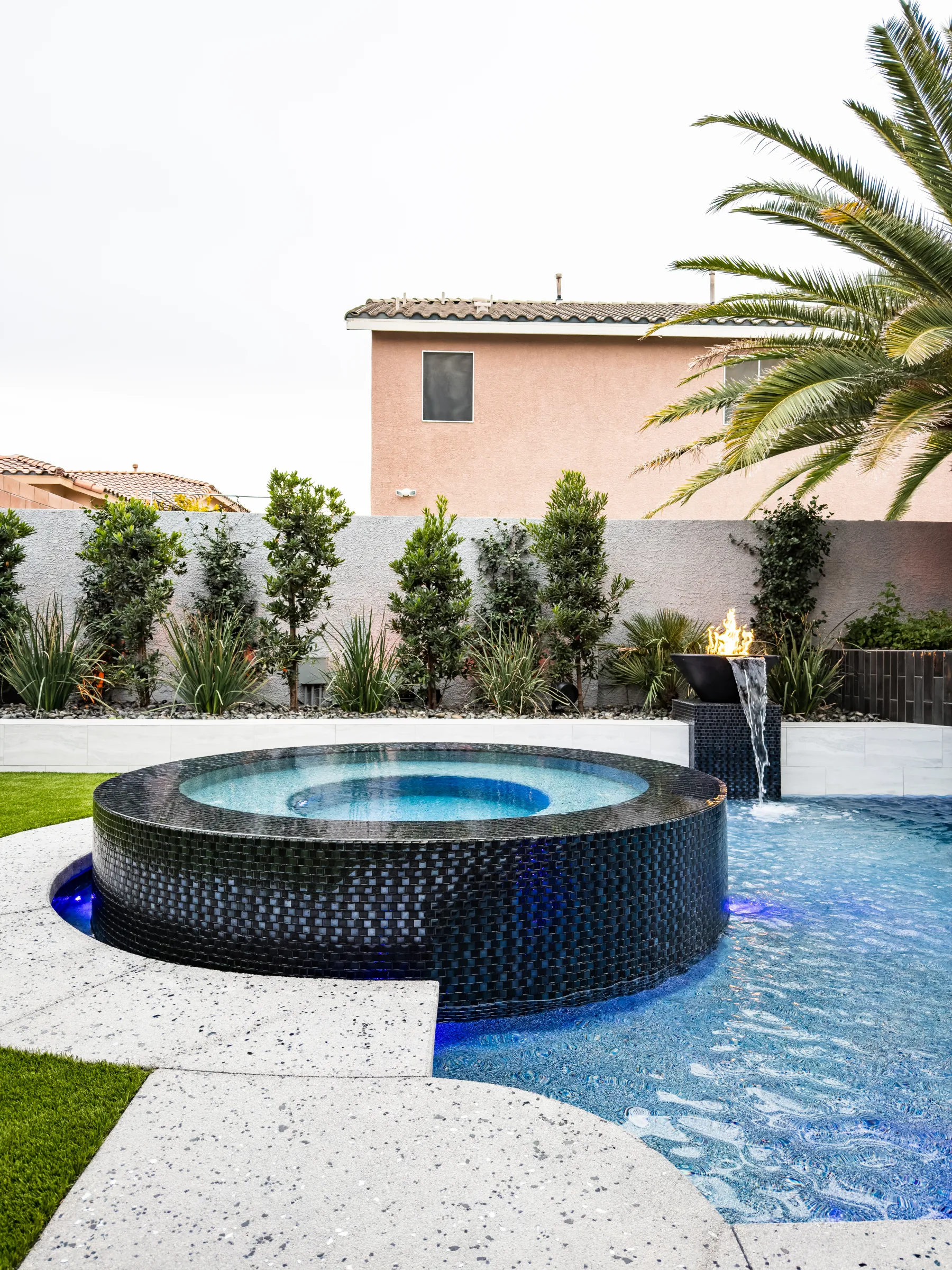 Round dark-tile spa with a fire bowl and a waterfall flowing into the pool