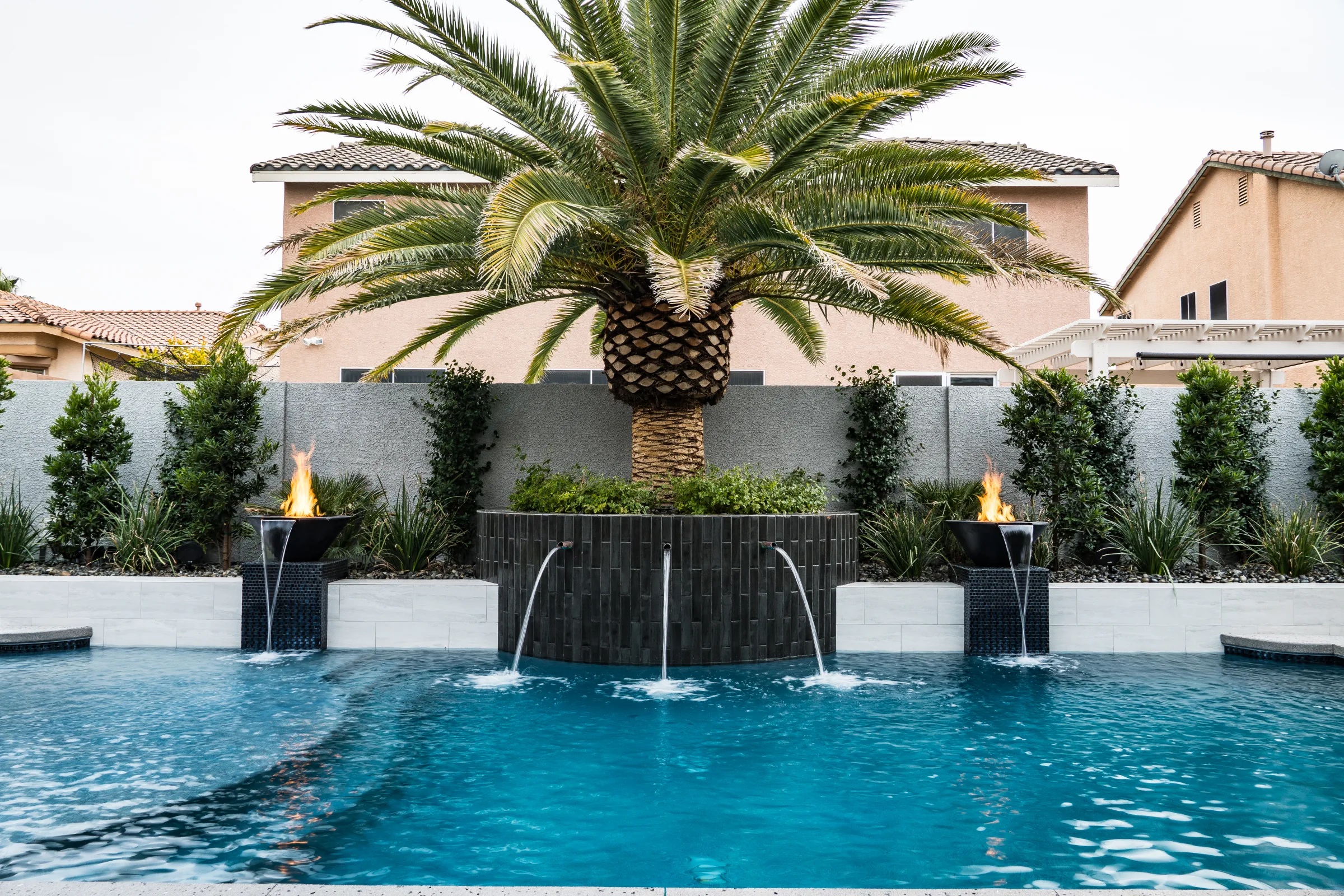 Wide backyard pool with waterfalls, fire bowls, palms, and a raised planter