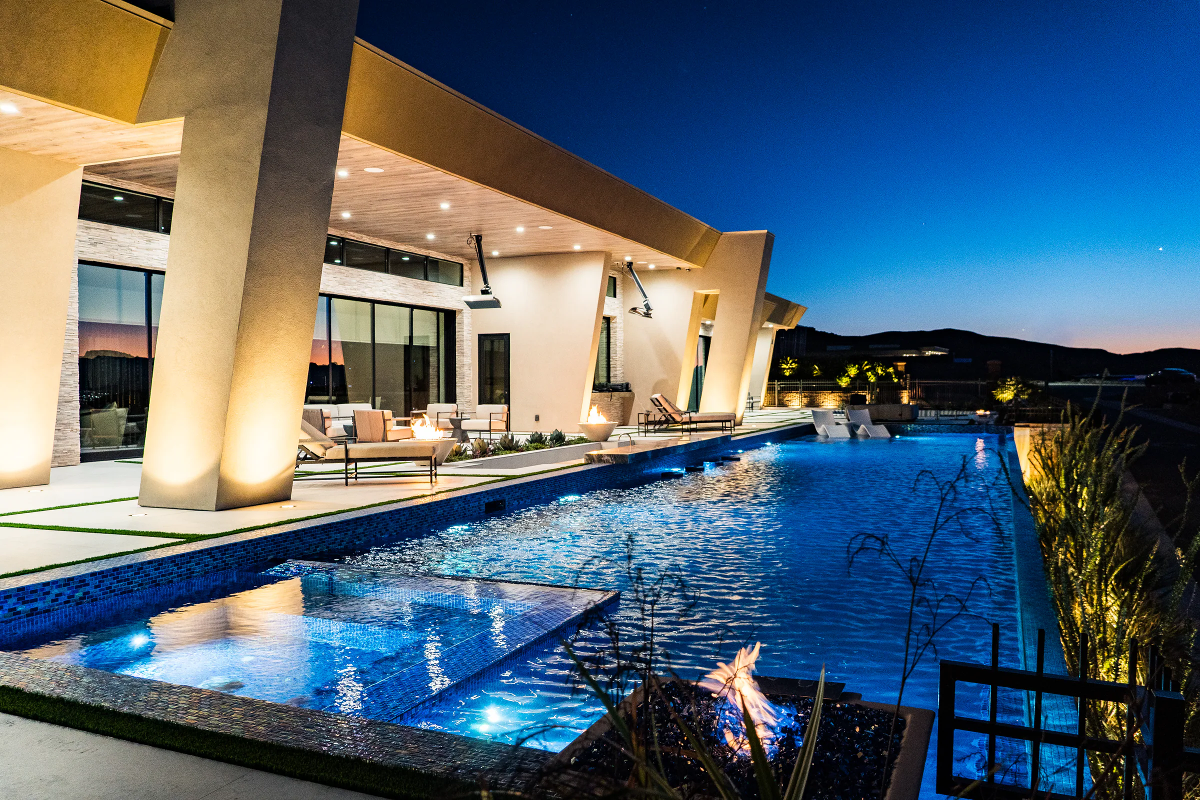 Lit infinity-edge pool, spa, and fire feature beside the covered patio at dusk