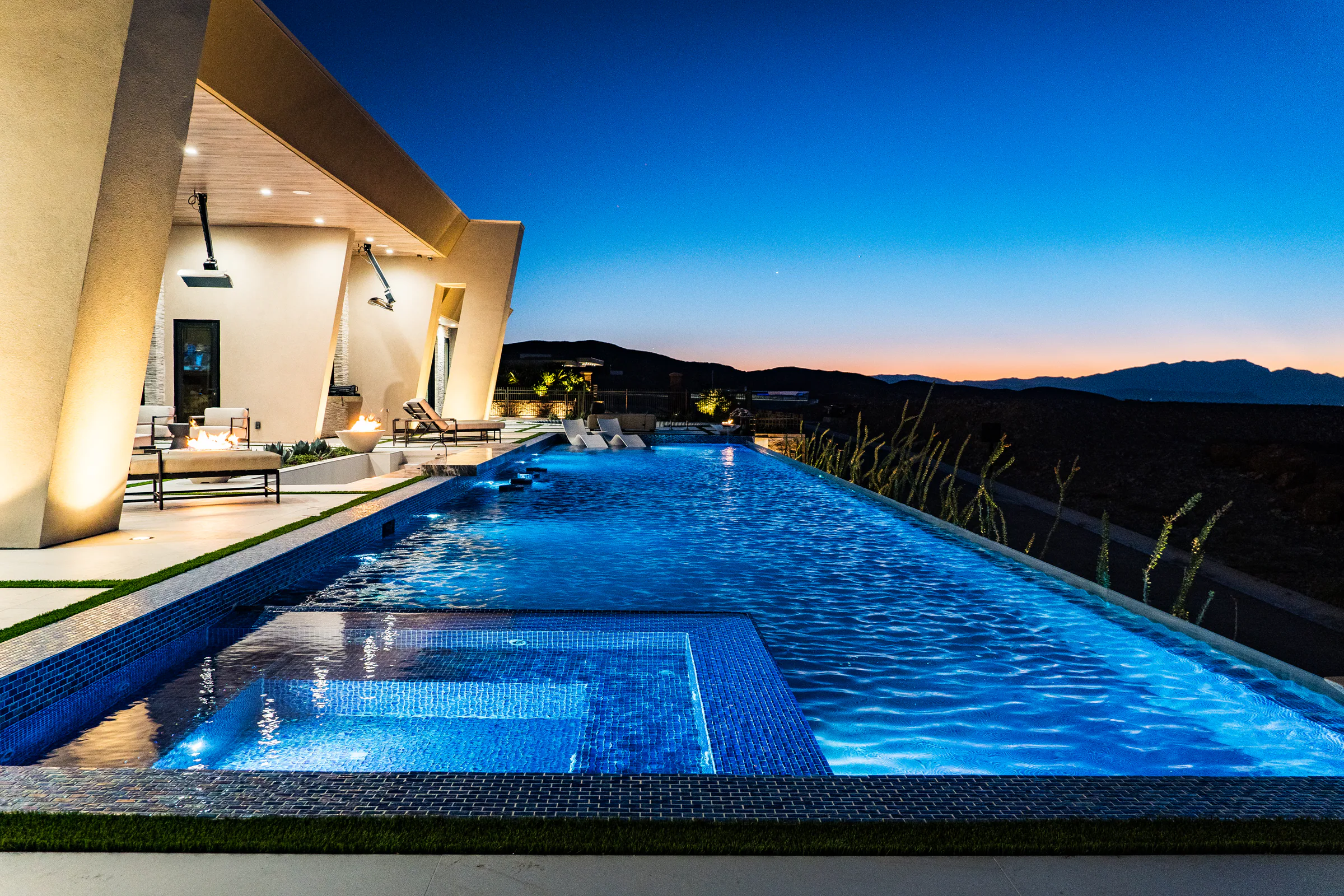 Infinity-edge pool and glass-tile spa glowing blue at dusk with valley lights beyond