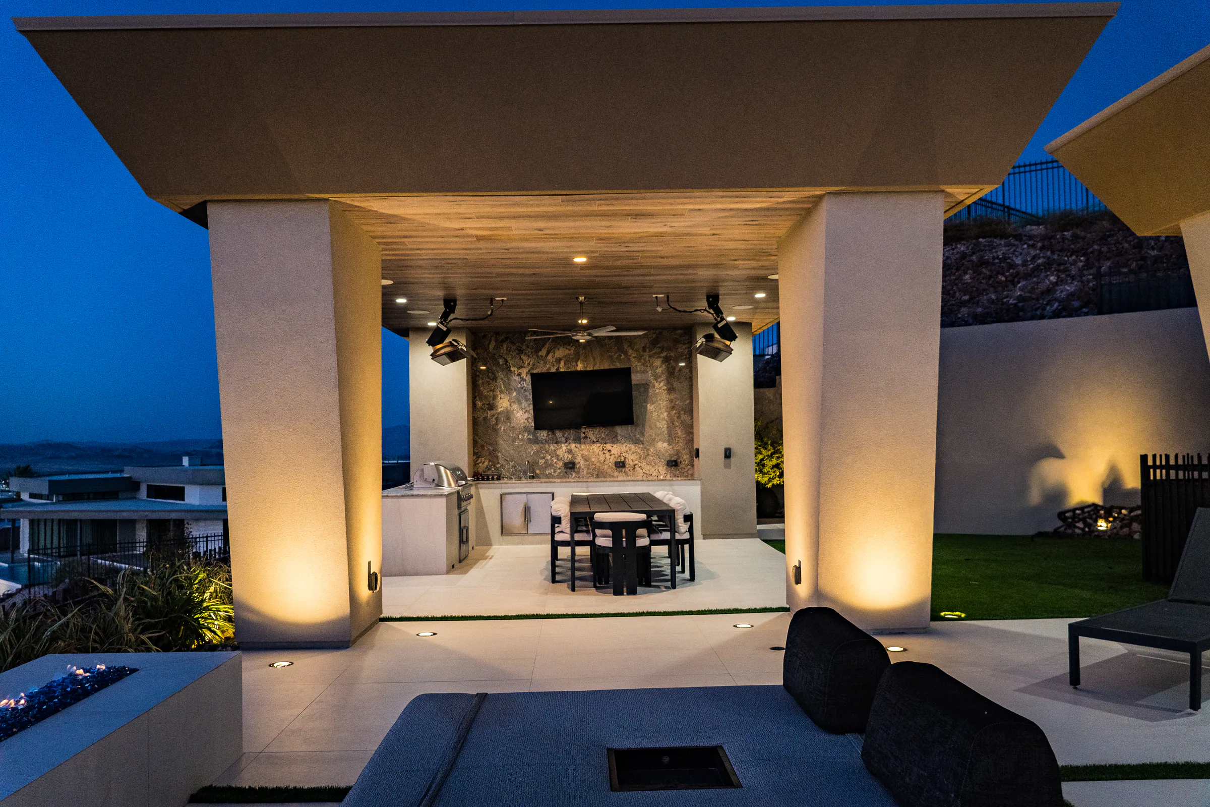 Covered pavilion with an outdoor kitchen, TV, and dining table behind a sunken lounge