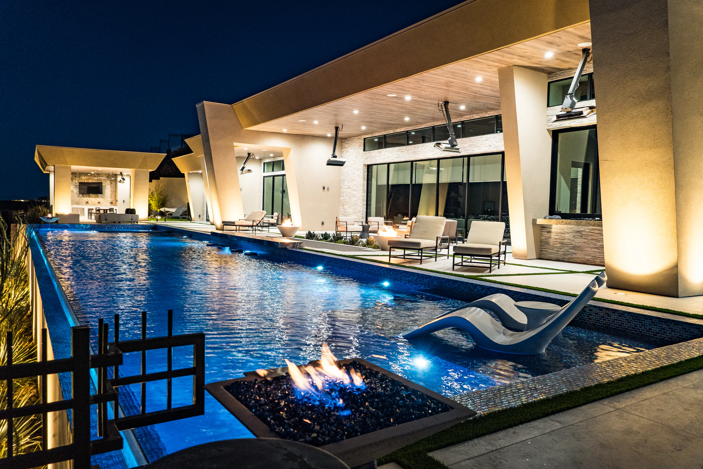Fire bowl and in-pool lounger beside the lit lap pool and covered patio at night