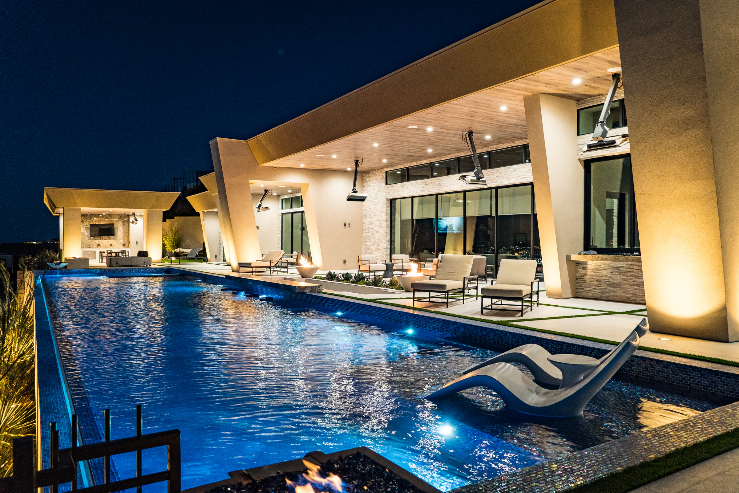 In-pool white loungers and a blue-lit lap pool along the home's covered patio